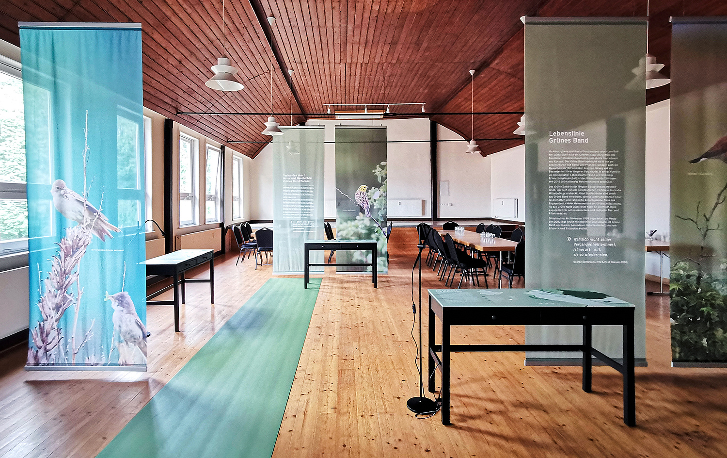 Das Bild zeigt einen Blick auf den Saal. Dieser ist mit von der Decke hängenden und im Design der Ausstellung gestalteten Stoffbannern gegliedert. (© Papenfuss | Atelier)
