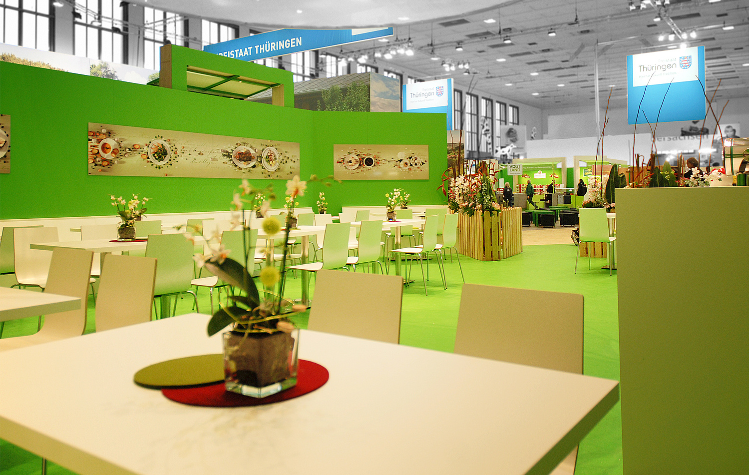 Blick vom VIP-Bereich mit mehreren Tischgruppen auf den Messestand. Die Tische und Stühle sind in weiß gehalten und mit grün-orangenen Tischsets dekoriert. (© Papenfuss | Atelier)