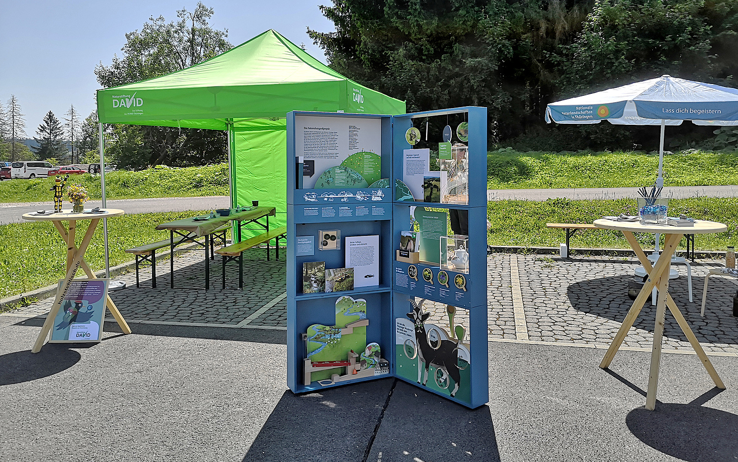 Aufgebautes Infomodul während einer Outdoorveranstaltung, im Hintergrund ein Pavillon ein Sonnenschirm und ein Tisch (© Papenfuss | Atelier)