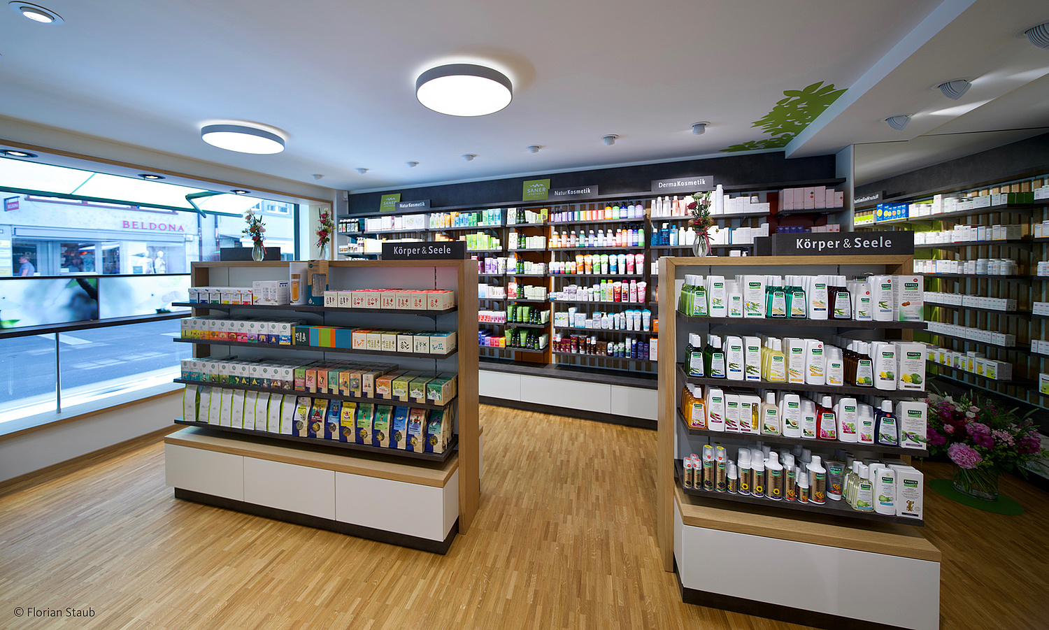 Innenarchitektur – Saner Apotheken – Store Liestal – Verkaufsbereich Blick in den Verkaufsbereich mit zwei freistehenden Einzelregalen und auf eine Wand mit Regal, jeweils mit Produkten befüllt (© Papenfuss | Atelier)